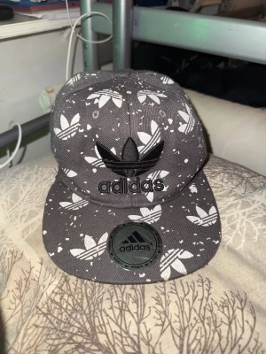 Svart Adidas keps med logomönster - Säljer en svart keps från Adidas med platt skärm och broderad logga framtill. Kepsen har ett vitt mönster med Adidas treklöver-loggor över hela ytan och små vita stänk. Justerbar baktill och tillverkad i bomull.