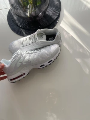 Nike Air Max Plus vita sneakers - Nike Air Max Plus i helvitt med mesh-överdel, ikoniska vågiga detaljer och synliga Air Max-enheter i sulan. Snygg design med grå och svarta detaljer på yttersulan och Tn Air-logga undertill. Perfekta för dig som vill ha en clean och sportig look.