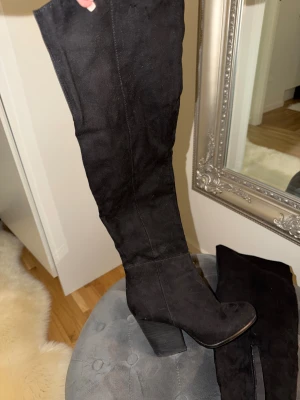 Svarta höga stövlar med klack - Snygga svarta stövlar i mocka med hög klack och rund tå. Stövlarna går över knäet och har dragkedja på insidan för enkel påtagning. Perfekta för att lyfta din outfit med en stilren och trendig look. 9 cm