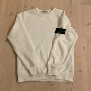 Stone island sweatshirt  - Vit sweatshirt från Stone Island med klassisk rund hals och ribbade muddar. Tröjan har den ikoniska svarta logotyp-patchen med gult och grönt broderi på vänster ärm. Tillverkad i mjuk bomull för en clean och stilren look.