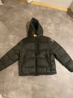 Mörk grön dunjacka Parajumpers Endurance - Säljer en svart dunjacka från Parajumpers, modell Endurance. Jackan har huva, dragkedja framtill och två sidofickor med dragkedja. Insidan är orange med extra ficka och coola detaljer. Perfekt för kalla dagar och riktigt snygg streetstil.