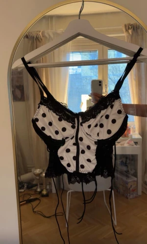 Svart och vit prickig korsett med spets - Säljer en snygg korsett-topp med svart spets och vita partier med svarta prickar. Toppen har smala axelband, snörning och dekorativa spetsdetaljer både fram och bak. Den är croppad och snörena som hänger kan man knyta till te.x rosetter och spänna så den sitter tajtare 