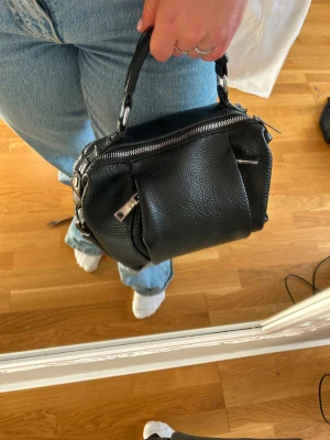 Svart handväska i skinn med dragkedja - Cool svart handväska i mjukt, strukturerat skinn med silverfärgade detaljer och flera dragkedjor. Väskan har ett robust handtag och praktiska fack både fram och bak. Perfekt storlek för att få med det viktigaste och ger en edgy vibe till din outfit. okänt märke