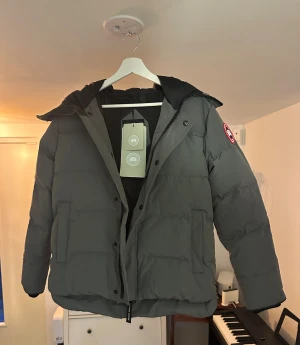 Canada Goose MacMillan  - Säljer en grå pufferjacka från Canada Goose med fluffig päls på huvan och klassisk logga på ärmen. Jackan har dragkedja, knappar och två stora fickor framtill. Perfekt för kalla dagar med sin tjocka och quiltade design. Canada Goose MacMillan grå med rött märke. 