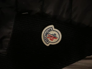 Svart Moncler cardigan med huva - Svart pufferjacka från Moncler med stickade ärmar och huva. Jackan har dragkedja framtill, Moncler-logga på vänster ärm och en ficka med dragkedja på ärmen. Kroppen är quiltad och tillverkad i syntetmaterial, medan ärmarna är stickade. Snygg och modern look.