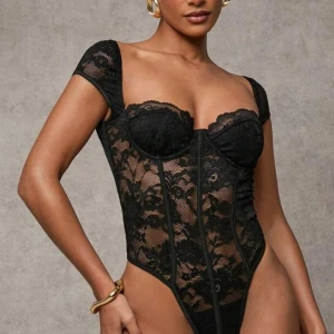 Svart spetsbody med kupor - Säljer en svart ”korsett”-body med genombruten spets och formade kupor. Den har korta ärmar och markerade sömmar framtill som ger en snygg siluett. Spetsen har blommigt mönster och materialet är mjukt och följsamt. Perfekt för dig som vill ha en statement piece.