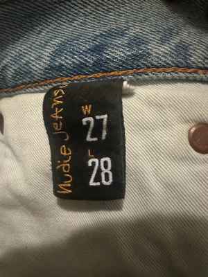 Ljusblå Nudie Jeans W27 L28 - Säljer ett par ljusblå jeans från Nudie Jeans med klassisk femficksdesign och snygga broderade detaljer på bakfickorna. Jeansen har rak passform och normal midja. Perfekt för dig som gillar en clean och tidlös look.