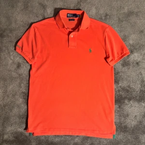 Pikétröja från Polo Ralph Lauren - Snygg korall/rosa färgad pikétröja från Polo Ralph Lauren med klassisk krage och två knappar framtill. Tröjan har grön broderad logga på bröstet och Polo-text på ärmen. Modellen är slim fit och har korta ärmar. Axelmåttet är 45 cm och längden är 68 cm.