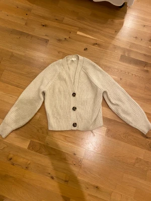 Beige ribbad kofta från H&M - Snygg beige kofta från H&M i storlek S. Modellen är ribbstickad med v-ringning och har tre stora knappar framtill. Koftan har långärmade ärmar och är gjord i mjukt stickat material som känns mjukt.
