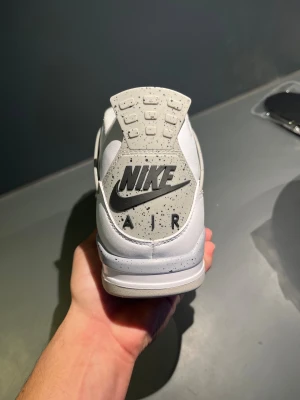 Nike Air Jordan 4 White Cement - Nike Air Jordan 4 White Cement med vit ovandel i skinn, grå och svarta detaljer samt specklad grå sula. Klassisk meshpanel på sidorna, svart Nike Air-logga på hälen och röd Jumpman på plösen. Snygg och ikonisk siluett som sticker ut.