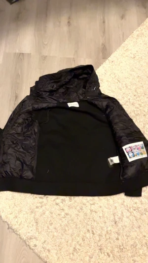 Svart Moncler pufferjacka med huva - Svart pufferjacka från Moncler med quiltad framsida och huva. Jackan har långärmade stickade ärmar, dragkedja framtill och Moncler-logga på ärmen. Perfekt för dig som gillar stilren design och vill ha något som håller värmen.