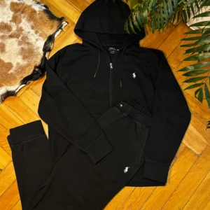 Svarta mjukisbyxor och zipper hoodie från Polo Ralph Lauren - Svarta mjukisbyxor från Polo Ralph Lauren med klassisk vit logga broderad på benet. Byxorna har ribbade muddar vid benslut, elastisk midja med snörning och mjukt bomullsmaterial som är skönt mot huden. Perfekt för chill dagar eller när du vill vara extra bekväm.