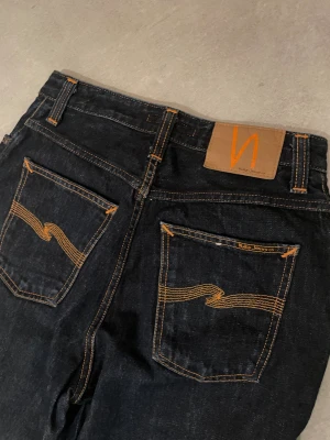 Nudie Jeans  - Snygga mörkblå Nudie Jeans Breezy Britt med klassisk orange söm och cool broderad detalj på bakfickorna. Modellen har hög midja och raka ben, tillverkade i 100% bomull för en autentisk jeanskänsla. Perfekt för dig som gillar stilrena och tidlösa jeans.