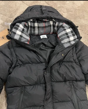 Svart pufferjacka från Burberry - Svart pufferjacka från Burberry med stor huva och klassiskt rutigt foder i grått, vitt och rött. Jackan har två dragkedjeförsedda fickor, ribbade muddar och en tydlig Burberry London England-patch på ärmen. Perfekt för kalla dagar med sin tjocka och quiltade design. Storlek L men passar båda M och S ta Köp inte den om du har Storlek L 