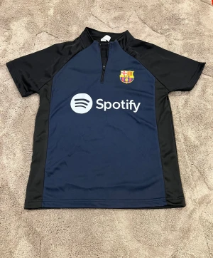 FC Barcelona tränings-tshirt med zip - Barn Storlek Snygg FC Barcelona tränings-tshirt med kort ärm, zip vid halsen och Spotify-logga på bröstet. Marinblå framtill med svarta ärmar och sidor, samt klubbmärke i färg. Tillverkad i lätt och ventilerande polyester, perfekt för träning eller match. Passar till barn från 9-11