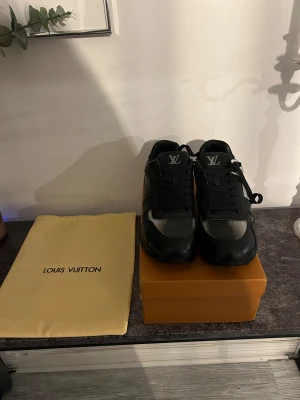 Louis Vuitton svarta och silver sneakers - Säljer ett par riktigt snygga Louis Vuitton sneakers i svart och silver med klassiskt LV-mönster. Skorna har rund tå, platt sula och är gjorda i skinn med detaljer i syntet. De har snörning och en stilren, modern look som sticker ut.