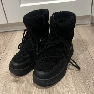 Svarta inuikii vinterskor men snörning  - PRIS GÅR ATT DISKUTERA.                                                   Inuikii i helsvart med fluffigt syntetmaterial och detaljer i skinn. Skorna har snörning runt hela skaftet. Perfekta för vintern och riktigt snygga till streetwear. Rund tå och högt skaft för maximal komfort. Lite nött på ovan sulorna, se bilder.  