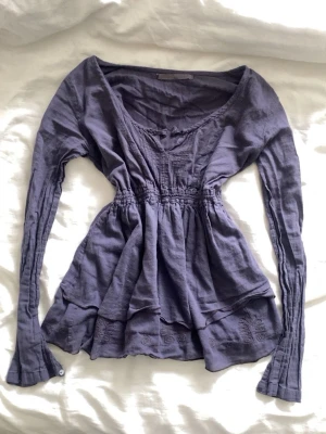 Lila blus med v-ringning och volang - Säljer en lila blus med v-ringning och långa ärmar. Blusen har smock i midjan för en snygg passform och volangdetaljer nedtill. Perfekt för dig som gillar en feminin och avslappnad stil.