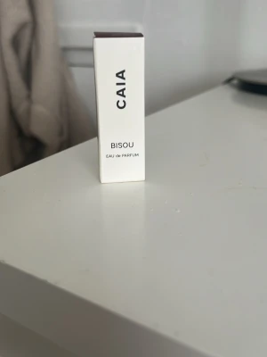 CAIA Bisou Eau de Parfum miniflaska - Säljer en liten parfymflaska från CAIA i doften Bisou. Flaskan är genomskinlig med vitt spraylock och kommer i en stilren vit och vinröd kartong. Perfekt att ha med i väskan för en fräsch doft när som helst.