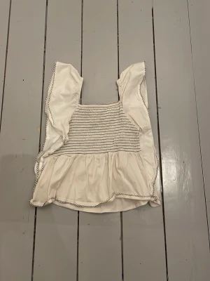 Vit volangblus med broderade detaljer - Söt vit blus med volangärmar och smockad överdel. Blusen har broderade kanter i kontrastfärg och en luftig, flowy passform. Perfekt för dig som gillar romantisk och bohemisk stil.