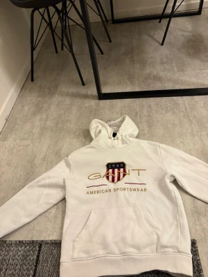 Vit hoodie från GANT med tryck - Snygg vit hoodie från GANT med broderad logga och amerikansk sköld på bröstet. Hoodien har huva med dragsko och en stor magficka. Tillverkad i mjukt bomullsmaterial som känns riktigt skönt mot huden. Perfekt för en avslappnad och sportig stil.
