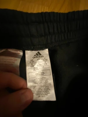 Adidas mjukisbyxor svart/grå XL - Snygga mjukisbyxor från Adidas i svart och grått med klassiska vita ränder längs sidorna. Byxorna har resår i midjan och muddar vid bensluten. Perfekta för chill eller träning, tillverkade i mjukt material för maximal komfort.