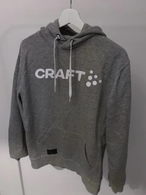 Grå hoodie från Craft med tryck - Snygg grå hoodie från Craft med vit logga framtill. Klassisk modell med huva och vita dragsnören samt en stor magficka. Perfekt för en avslappnad och sportig stil. Mjuk bomullsblandning som känns skön mot huden.