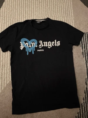 Palm Angels svart t-shirt  - Säljer en svart t-shirt från Palm Angels med stort tryck framtill. T-shirten har ett blått droppande hjärta och texten 'Palm Angels Paris' i vitt. Klassisk passform och rund hals. Perfekt för dig som gillar streetwear och vill sticka ut.