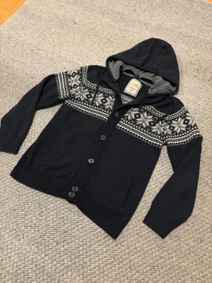 Mörkblå kofta med mönster och luva - Mysig mörkblå stickad kofta med vit snöflingemönster över bröst och axlar. Koftan har luva, knäppning med knappar framtill och ribbade muddar vid ärmslut och nederkant. Perfekt för kalla dagar och ger en avslappnad stil. Storlek M. Luvan går o ta bort om man skulle vilja de.