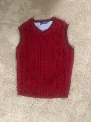 Ralph Lauren väst! - Snygg röd kabelstickad väst från Ralph Lauren med V-ringning och mörkblå detaljer vid hals och ärmöppningar. Klassisk broderad logga på bröstet. Perfekt att bära över skjorta för en skön look. Storlek XL i barn men passar som en S i man eller M kvinna! Ny pris är 1200kr. Priset är rörligt!