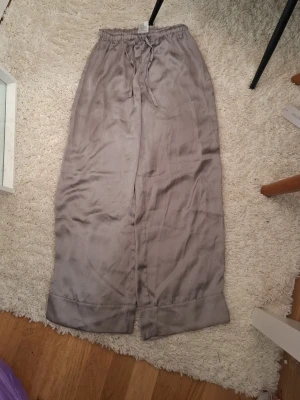 Grå loungebyxor med snörning - Säljer ett par grå loungebyxor med sidenliknande känsla med avslappnad passform och breda ben. Byxorna har resår i midjan och snörning. Hög midja men ser även bra om man har de lågt. Säljer för att de inte används längre, är även inte mycket använda.