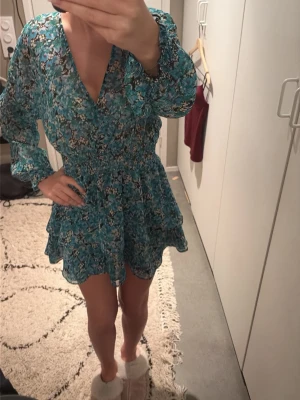 Blommig blå klänning från Zara - Supersöt blommig klänning från Zara i blå och gröna toner. Klänningen har v-ringning, långa ärmar med resår vid handlederna och volangdetaljer vid kjolen. Kort modell med smock i midjan för en snygg passform. Perfekt för vår och sommar!