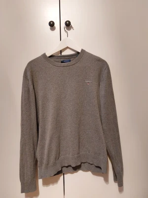 Grå stickad tröja från GANT - Snygg grå stickad tröja från GANT med rund halsringning och diskret logga broderad på bröstet. Tröjan har långa ärmar och ribbade muddar vid ärmslut och nederkant. Perfekt för lager på lager och enkel att matcha med olika stilar. Jag är stark under 180 cm och väger omkring 65kg
