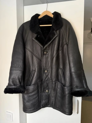 Svart shearlingjacka med knappar - Säljer en svart shearlingjacka av 100% skinn/shearling med bred krage och stora fickor framtill. Jackan har knäppning med stora knappar och mjuk päls på insidan samt vid ärmsluten (går att vika ner). Perfekt för kalla dagar och ger en cool, oversized look.