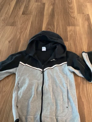 Grå och svart hoodie med dragkedja - Snygg hoodie i grått och svart med dragkedja framtill och huva. Hoodien har en vit detalj över bröstet och svarta partier på axlar och ärmar. Perfekt för en avslappnad streetwear-look.