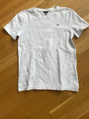 Vit t-shirt från Tommy Hilfiger - Vit t-shirt från Tommy Hilfiger med liten logga på bröstet och en vertikal röd, vit och marinblå rand samt TH-tryck på ryggen. Klassisk passform och rund halsringning. Perfekt till jeans eller shorts.