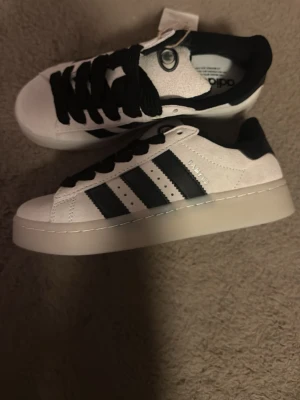 Adidas Campus sneakers vitbeige/svart - Stilrena Adidas Campus sneakers i vitt/beige mocka med svarta detaljer och klassiska tre ränder på sidan. Skorna har svart snörning, rund tå och platt sula. Perfekta för dig som gillar en clean och tidlös look.