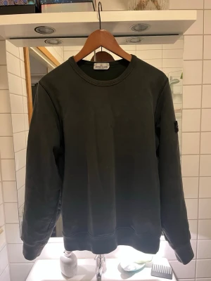 Mörkgrå sweatshirt från Stone Island - Snygg mörkgrå sweatshirt från Stone Island med klassisk rund halsringning och ikonisk logotyp-patch på ärmen. Tröjan har en avslappnad passform och ribbade muddar vid ärmslut och nederkant. Perfekt för en stilren och trendig look. Passar både till mer uppklätt med en skjorta under men också till skolan.