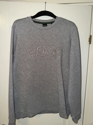 Grå tröja från BOSS - Snygg grå sweatshirt från BOSS med broderad logga framtill. Tröjan har rund halsringning, långa ärmar och är gjord i mjukt material som känns skönt mot huden. Perfekt för en avslappnad och stilren look.