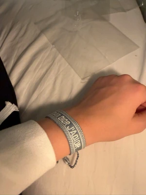 Dior armband  - Snyggt grått armband från Christian Dior med broderad logga och justerbar knytning med tofsar. Armbandet är gjort i textil och har en trendig, avslappnad look som passar perfekt till din outfit.