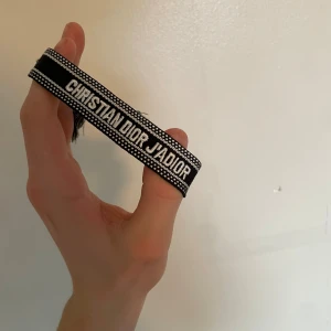 Christian Dior J'Adior tygarmband - Snyggt svart och vitt tygarmband från Christian Dior med texten 'J'ADIOR' broderad. Armbandet har en trendig och ungdomlig design med justerbar knytning och små tofsar i ändarna. Perfekt accessoar för att lyfta din outfit.