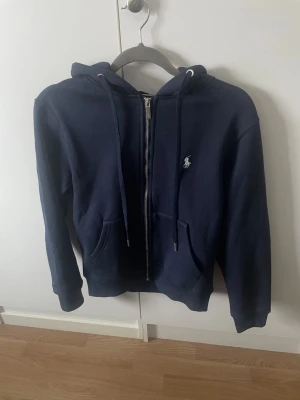 Mörkblå hoodie från Polo Ralph Lauren - Snygg mörkblå hoodie med dragkedja från Polo Ralph Lauren. Klassisk design med huva, dragsnören och två fickor framtill. Ikoniska loggan broderad på bröstet. Tillverkad i mjukt bomullsmaterial som är skönt att ha på sig.Och varm