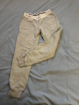 Grå mjukisbyxor från Calvin Klein - Snygga grå mjukisbyxor från Calvin Klein med bred resår i midjan där loggan syns tydligt. Byxorna har raka ben, två framfickor och ribbade muddar vid bensluten. Perfekta för chill eller träning.