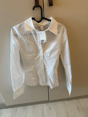 Vit klassisk skjorta från H&M - Klassisk vit skjorta från H&M med lång ärm och krage. Skjortan har knappar framtill och är tillverkad i bomull, perfekt för en clean och stilren look. Passar till allt från jeans till kostymbyxor. Som en xs
