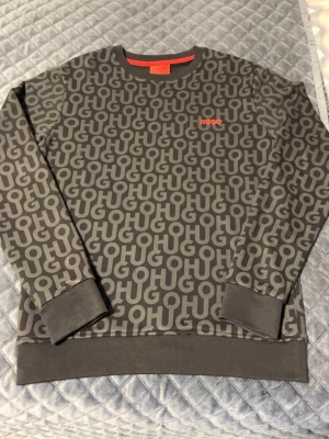 Grå sweatshirt från HUGO med logotyp - Snygg grå sweatshirt från HUGO med all-over logotypmönster i ljusgrått och röd HUGO-text på bröstet. Tröjan har rund hals, ribbade muddar och nederkant. Perfekt för dig som gillar streetwear och vill sticka ut med en trendig look.