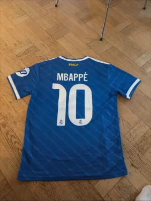 Blå Real Madrid fotbollströja Mbappé - Säljer en blå Real Madrid fotbollströja från Adidas med Mbappé och nummer 10 på ryggen. Tröjan har vita detaljer, klubbmärke på bröstet och klassiska Adidas-ränder på axlarna. Materialet är lätt polyester och perfekt för fotbollsträning.
