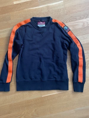 Svart sweatshirt från Parajumpers - Cool svart sweatshirt från Parajumpers med orangea ränder längs ärmarna och ett broderat PJS-märke på axeln. Tröjan har rund halsringning, ribbade muddar och är tillverkad i mjuk fleece. Perfekt för en avslappnad streetwear-look.
