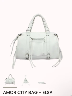 Vit Amor City handväska Elsa - Snygg vit handväska från Amor, modell City Bag Elsa. Väskan har flätade handtag, dekorativa dragkedjor och långa remmar på sidorna. Tillverkad i syntetmaterial och har en stilren, modern look som passar till många outfits. Den är även helt oanvänd med lappar och plast kvar.