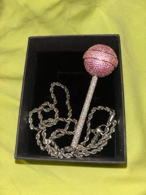 Halsband med lollipop-hänge - Unikt halsband med ett stort lollipop-hänge täckt av rosa och vita glittrande stenar. Hänget har formen av en klubba med en rund boll och lång pinne, båda dekorerade med gnistrande stenar. Kedjan är i silverfärgad metall med tvinnad design. Perfekt statement piece för dig som gillar att sticka ut.