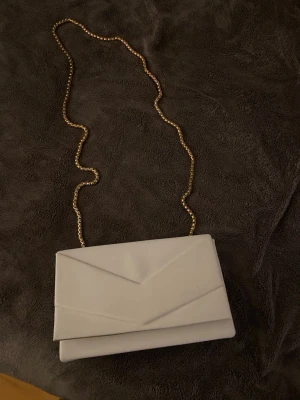 Vit axelväska med guldkedja - Snygg vit axelväska i kuvertmodell med guldfärgad kedja som axelrem. Väskan har en stilren och minimalistisk design med vikta detaljer på framsidan. Perfekt för dig som vill ha en enkel men elegant accessoar.